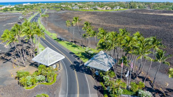 Aerial view of a coastal road winding through tropical landscaping with palm trees, two small pavilions, and volcanic lava fields nearby.