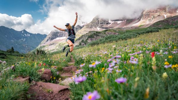A person joyfully jumps on a trail surrounded by colorful wildflowers, with mountains and clouds in the background.
