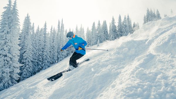A skier in a blue jacket descends a snowy slope surrounded by tall, snowy trees under a bright sky. The scene is wintery and vibrant.
