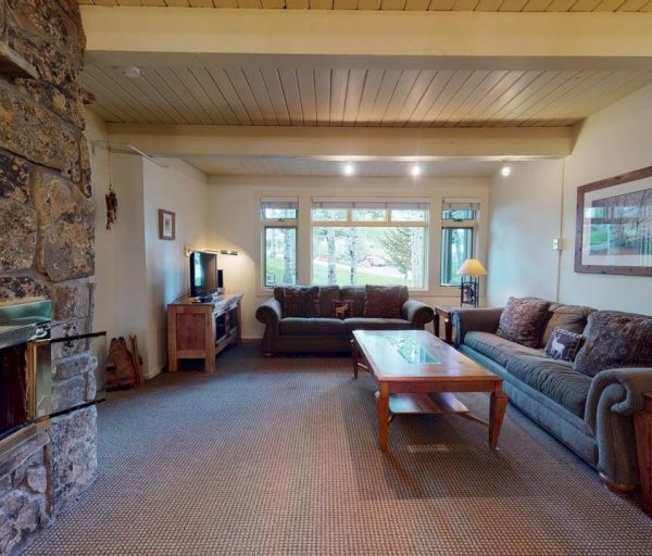 The image shows a cozy living room with a stone fireplace, comfy couches, a coffee table, and large windows letting in natural light.