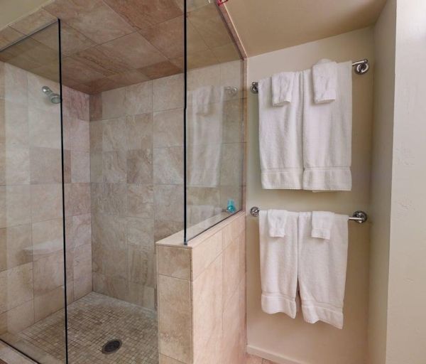 The image shows a clean bathroom with a glass shower, stone tiles, and hanging white towels. It has a simple and modern design.