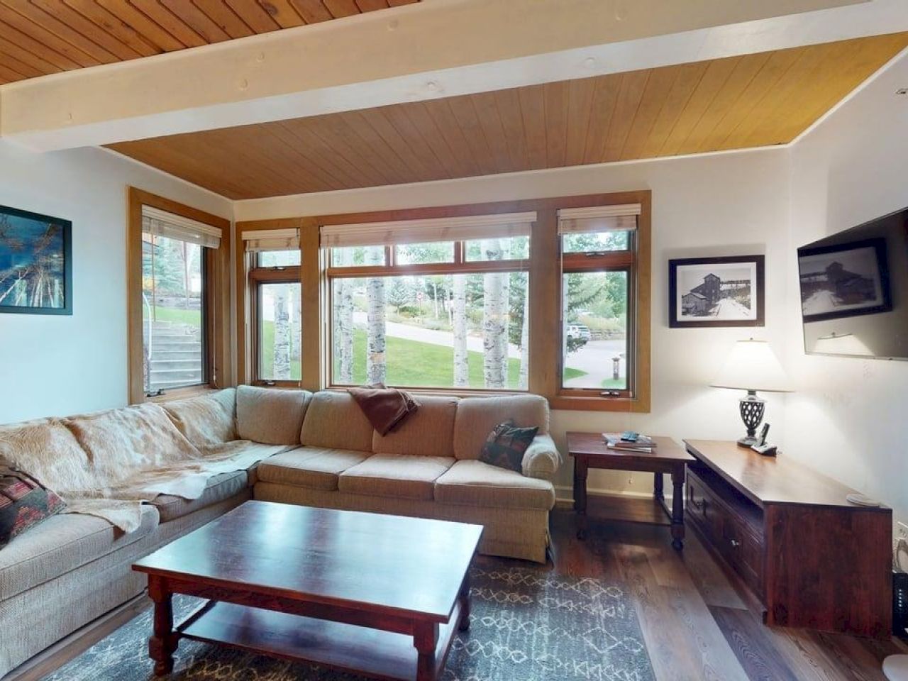 The image shows a cozy living room with a sectional sofa, coffee table, and large windows overlooking a green area outside.