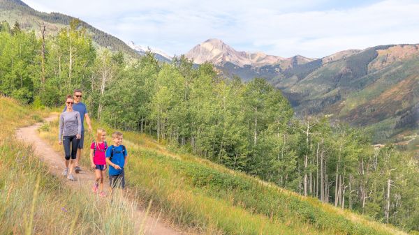 A family walks along a trail through green trees and mountains, enjoying a scenic day outdoors together.
