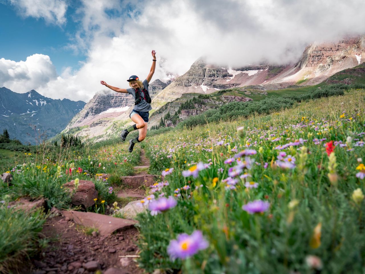 A person joyfully jumps on a mountain trail surrounded by vibrant wildflowers and stunning mountainous scenery.