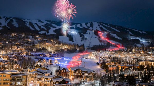 The image showcases a snowy mountain landscape at night, illuminated by fireworks and colorful ski trails, creating a festive atmosphere.