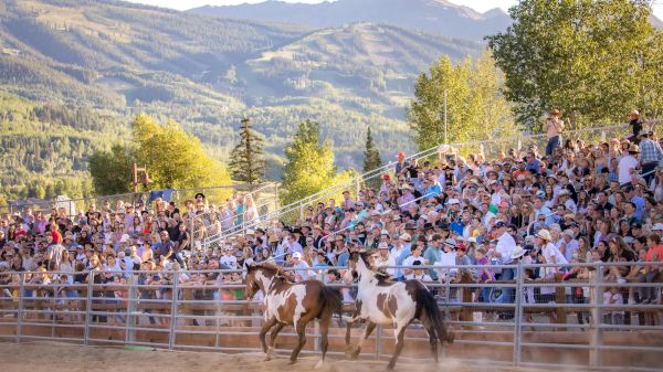 The image features a rodeo scene with horses running and a large crowd of spectators against a mountain backdrop.