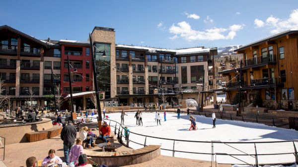 The image features a bustling outdoor ice rink surrounded by modern buildings, with people enjoying skating and dining nearby.