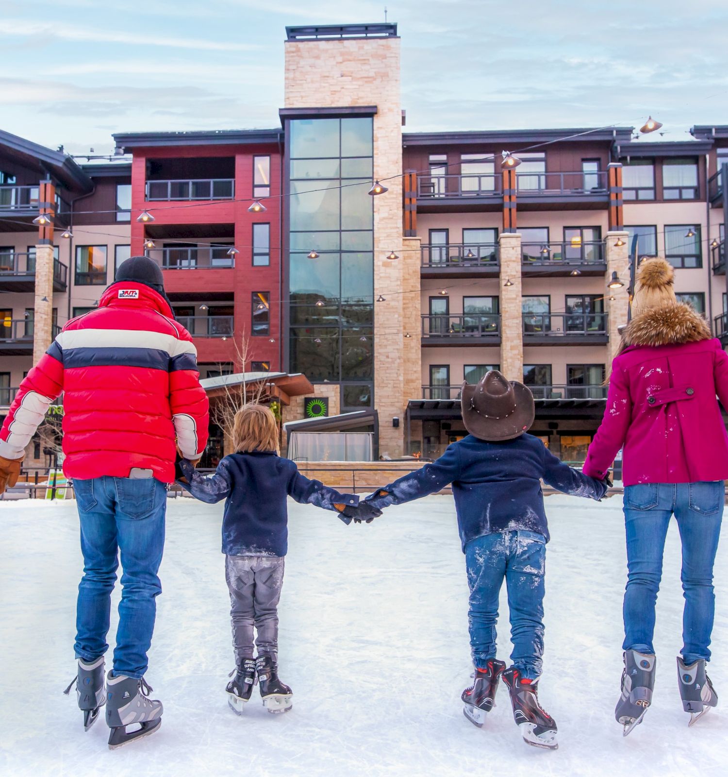 A family skating on an ice rink outside a modern building, enjoying time together while holding hands and wearing winter attire.