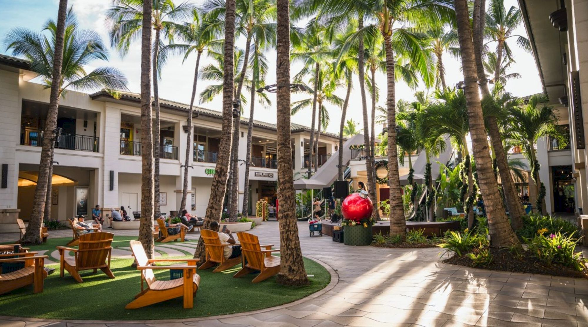 The image shows a lively outdoor space with palm trees, adirondack chairs, shops, and people enjoying the sunny atmosphere.