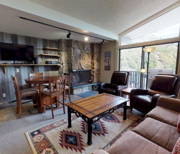 The image shows a cozy living room with rustic decor, a dining table, plush seating, a fireplace, and large windows offering a view.