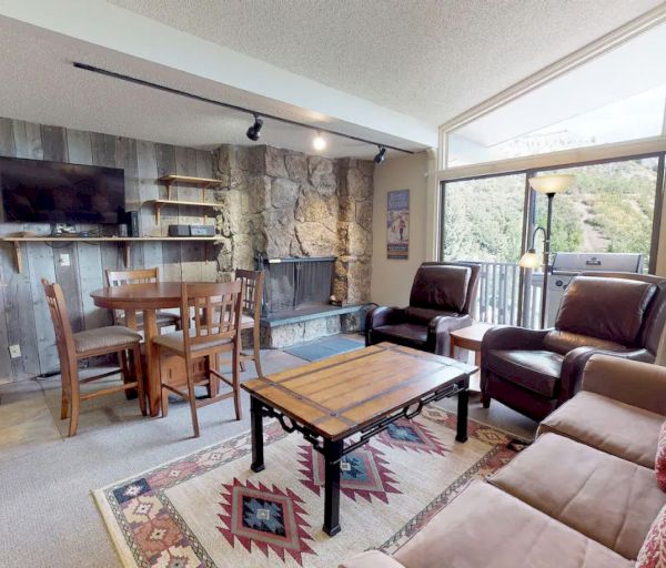 The image shows a cozy living space with rustic decor, a stone fireplace, a dining area, and large windows showcasing nature.