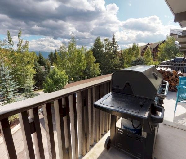 The image shows a balcony with a grill, turquoise chairs, and greenery all around, set against a scenic cloudy backdrop.