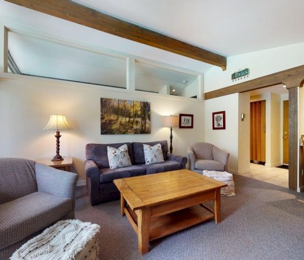 The image shows a cozy living area with comfortable couches, a wooden coffee table, and large windows letting in natural light.