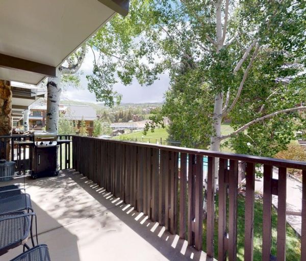 The image shows a balcony with two chairs, a grill, and trees, overlooking a green lawn and scenic view. It's a peaceful setting.