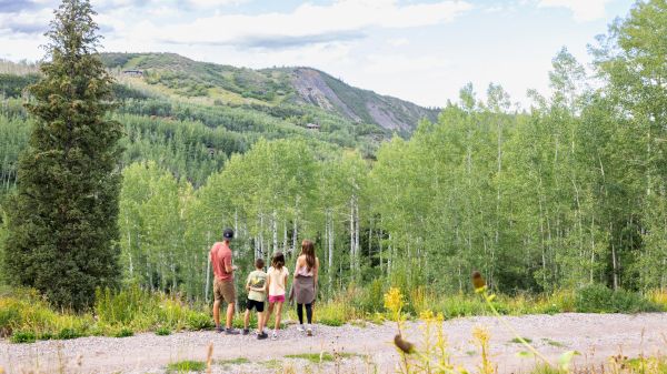 A family stands on a path, gazing at a lush green landscape with trees and mountains under a partly cloudy sky.