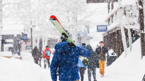 A person in a blue jacket carries a snowboard over their shoulder on a snowy street, others walking by in winter gear.