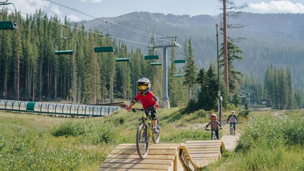 Children biking on a wooden path in a forested area, with chair lifts in the background under a clear sky. It&rsquo;s an outdoor adventure.