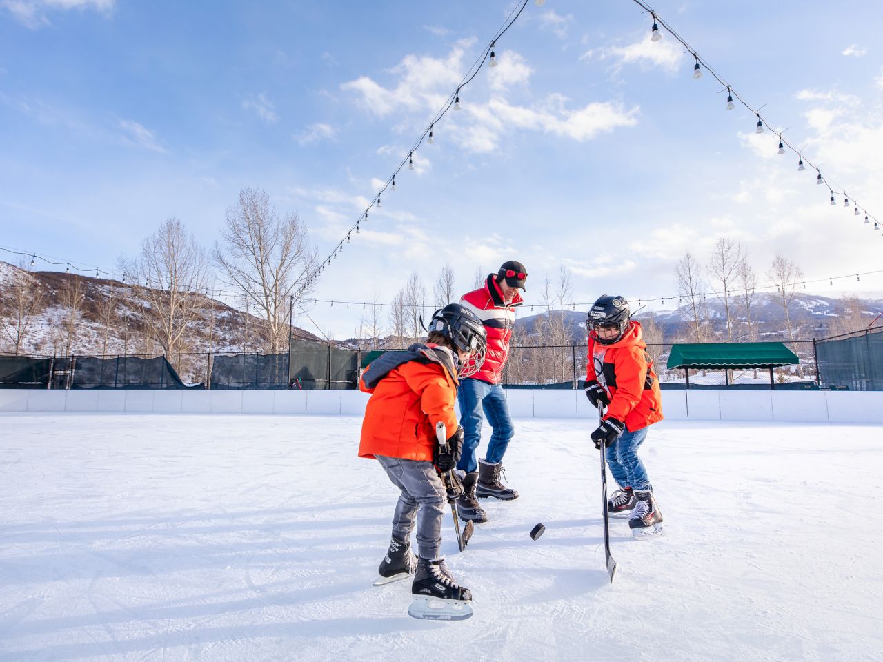 A coach guides kids on an ice rink, all wearing orange jackets while playing hockey under a clear blue sky.
