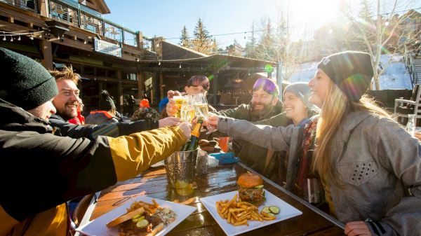 A group celebrates with cheers at a table, enjoying food and drinks in a sunny outdoor setting, surrounded by snowy scenery.