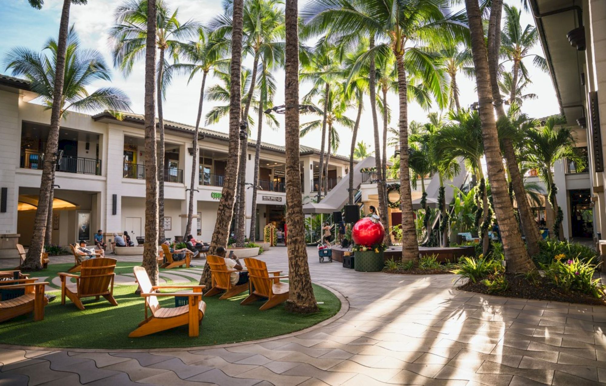 The image features a vibrant courtyard with palm trees, seating areas, and people, evoking a relaxed tropical atmosphere.