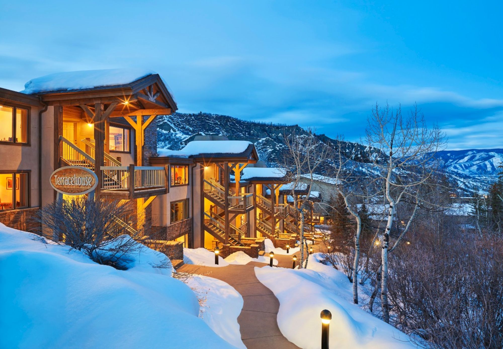 A cozy mountain lodge nestled in deep snow, warm lights glowing as dusk settles over snowy slopes and bare trees. (end)