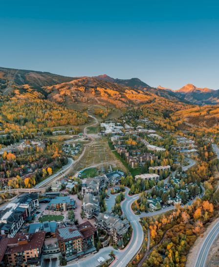 Aerial view of a picturesque mountain town with colorful autumn foliage, winding roads, and modern buildings beneath a clear sky.