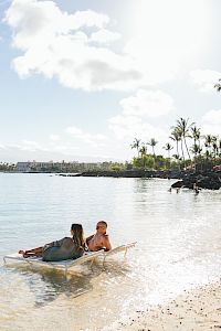 A couple relaxes in shallow water on a beach, with palm trees and a clear sky in the background. Perfect for a vacation vibe.
