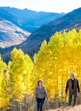 A couple enjoys a walk through vibrant yellow foliage in a mountainous landscape, capturing the beauty of autumn nature.