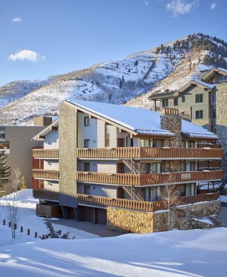 A snowy landscape features a cozy multifamily building surrounded by mountains and trees, creating a serene winter atmosphere.