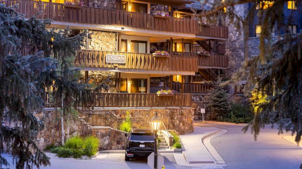 The image shows a mountain lodge with stone walls, balconies adorned with flowers, and a black vehicle parked outside.