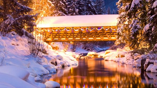 A snowy landscape features a lit wooden bridge over a calm river reflecting colorful lights and surrounded by tall evergreen trees.
