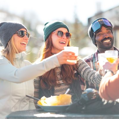 A group of friends enjoying drinks and food outdoors, smiling and celebrating in a sunny, wintery setting.