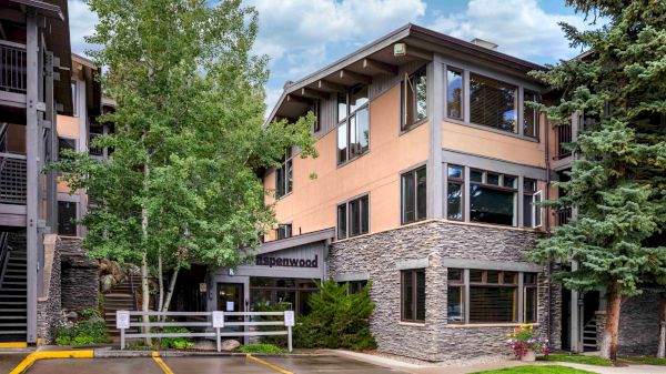 The image shows a modern building entrance with stone accents, large windows, and surrounding greenery. It's inviting and well-maintained.
