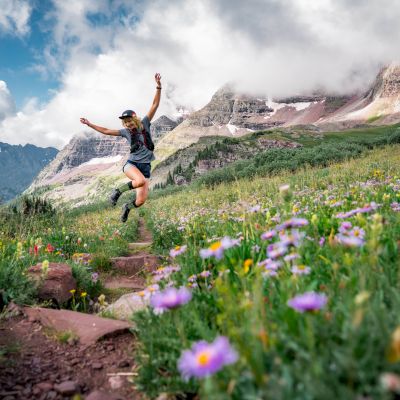 A person joyfully jumps along a trail surrounded by vibrant wildflowers and stunning mountainous scenery under a cloudy sky.