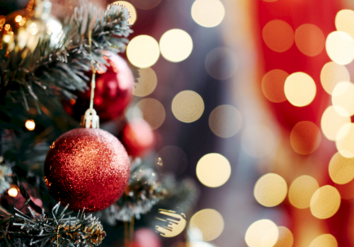 A decorated Christmas tree with red baubles and warm, blurred holiday lights in the background.