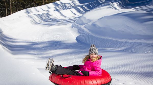A child in a pink jacket rides a red snow tube down a snowy slope, trees in the background, icy tracks ahead.