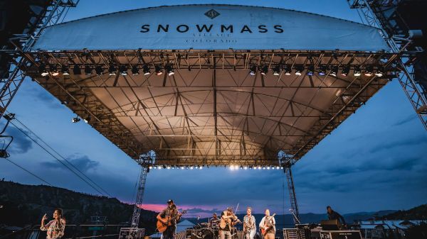 A live band performs on a large outdoor stage labeled &ldquo;Snowmass&rdquo; at dusk, with lights and rigging, mountains in the background.