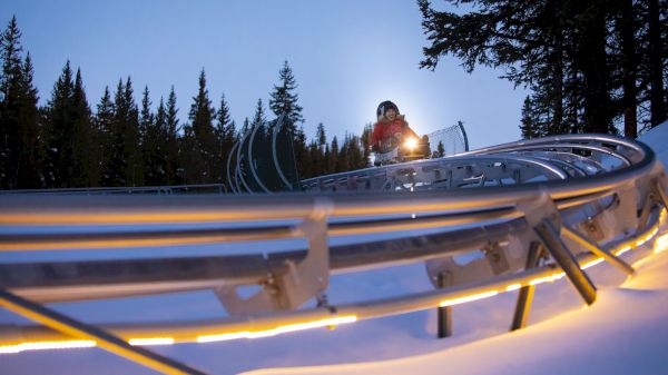 A person with a headlamp atop a snowy ski slope, railings and trees in the background at dusk, ready for night skiing.