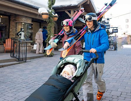 Two people wearing helmets walk a baby in a stroller, each carrying skis on their shoulders, along a street with shops.
