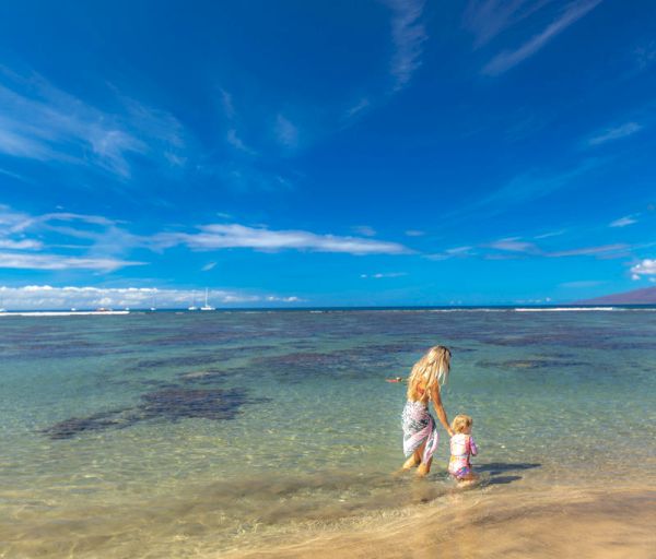A woman and a child wading in clear tropical water at the beach, with blue sky, gentle waves, and sandy shore.