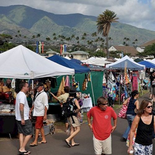 A bustling outdoor market with white tents, vendors selling goods, shoppers strolling, and a mountain backdrop under a sunny sky. (ends with period)