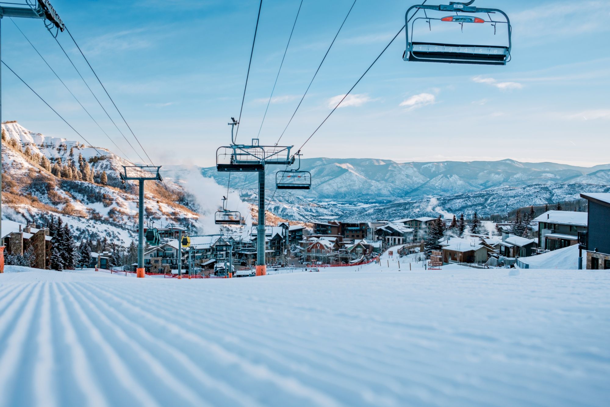 A snowy ski resort with chairlifts, empty groomed slopes, and distant mountains under a clear blue sky.