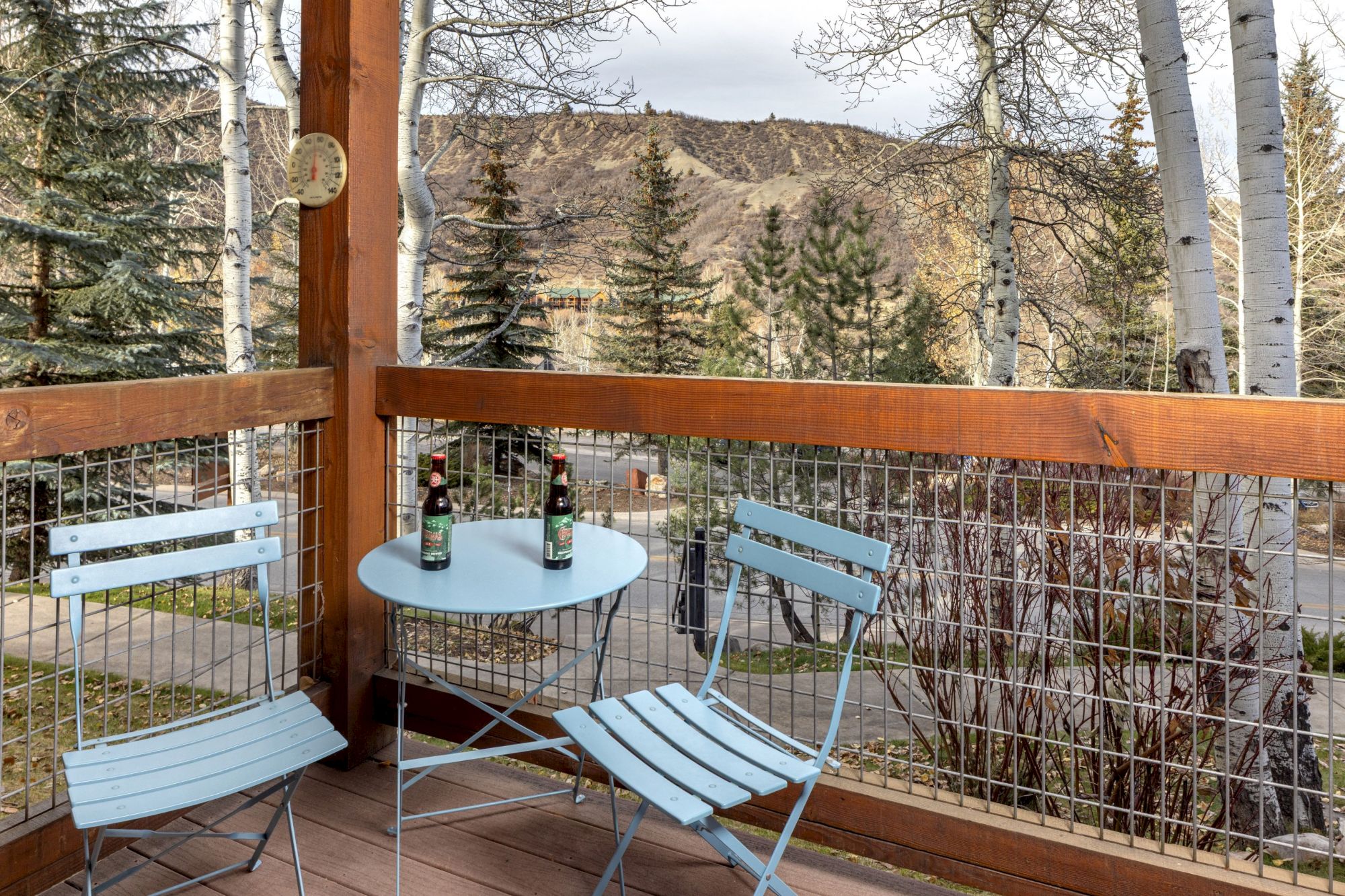 A small blue metal table and two matching chairs on a wooden balcony, with bottles on the table, trees and mountains in the background.