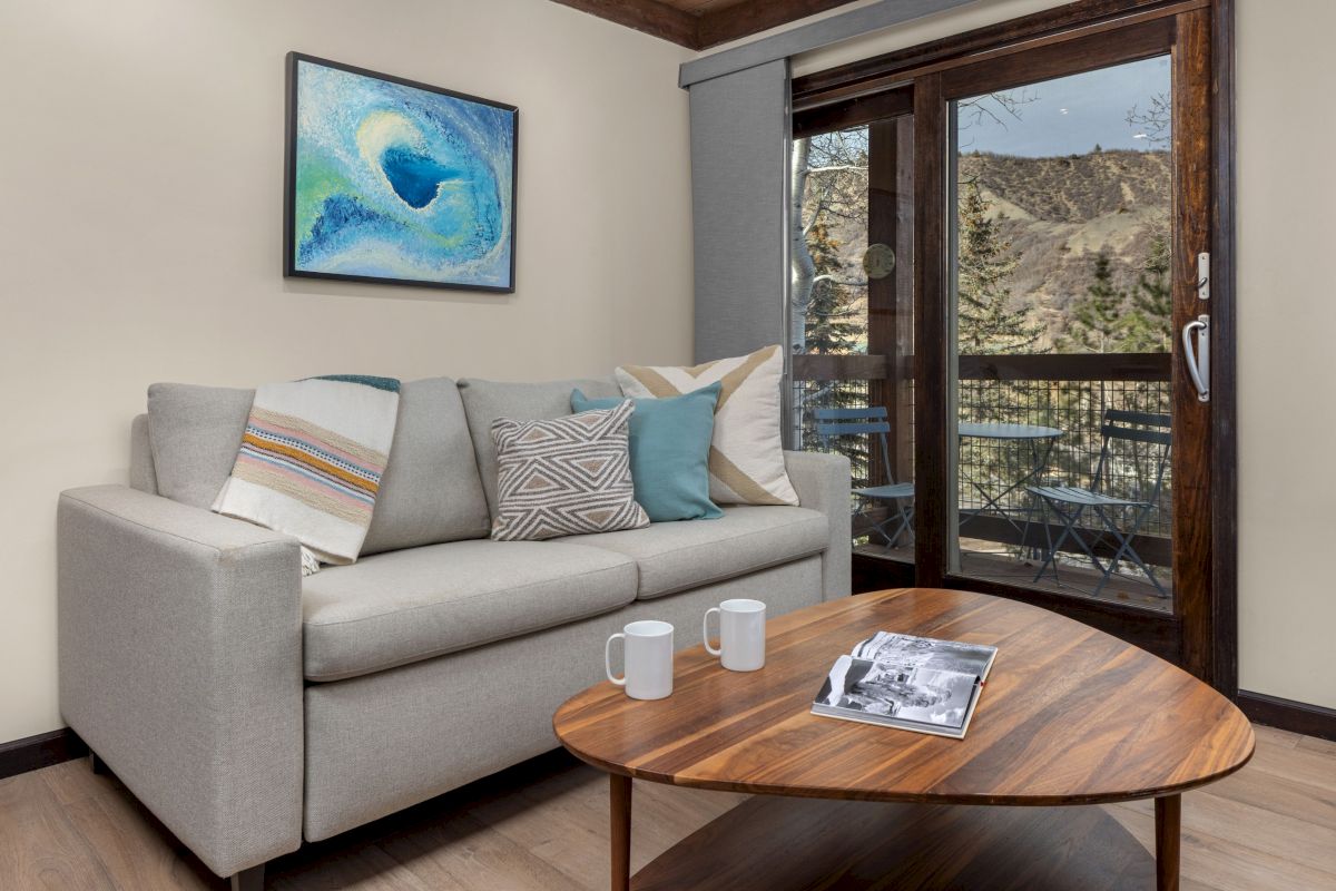 A cozy living room with a gray sofa, colorful throw pillows, a wooden coffee table, a framed art piece, and a balcony door showing a mountain view.