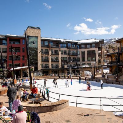 A bustling outdoor ice rink surrounded by modern apartments, with skaters, spectators, and sunny blue skies, in a lively plaza setting.