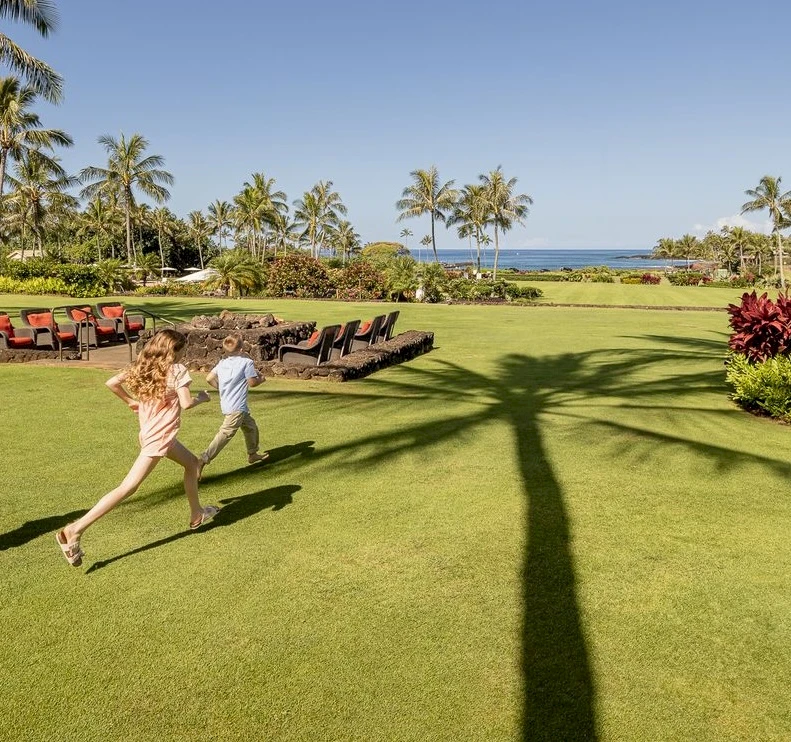 Two people sprint across a sunny, tropical lawn with palm trees, lounge chairs, and the ocean in the background, casting long shadows.
