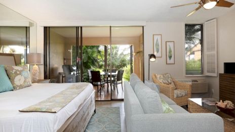 A bright hotel suite with a king bed, seating area, sliding glass doors to a balcony/patio, and tropical decor.