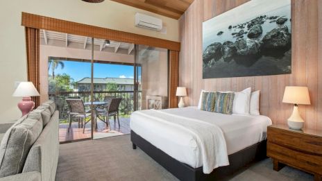 A cozy hotel room with a king bed, nightstands, sofa, balcony, and a sliding door to an outdoor seating area overlooking greenery and trees.