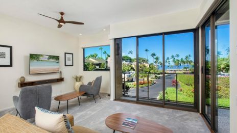 A modern living room with large floor-to-ceiling windows, a wooden coffee table, a gray sofa, a small desk and chairs, tropical landscape outside, and a ceiling fan.