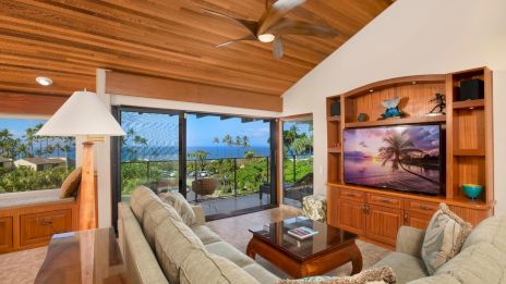 A bright coastal living room with a vaulted wooden ceiling, large windows to a tropical view, beige sofas, a wooden entertainment wall, and a glass sliding door.
